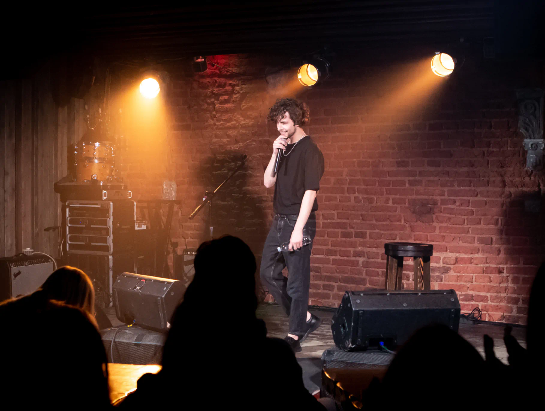 Interior of a dark comedy club with a man on the stage holding a mic in front of a brick wall. Bight lights shine on the stage.