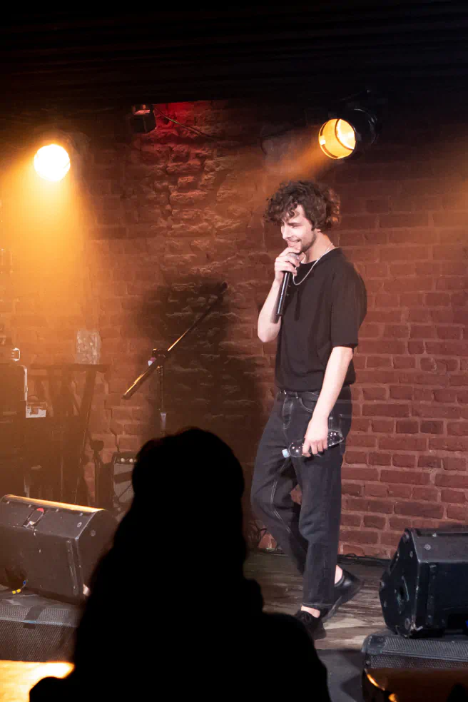 Interior of a dark comedy club with a man on the stage holding a mic in front of a brick wall. Bight lights shine on the stage.