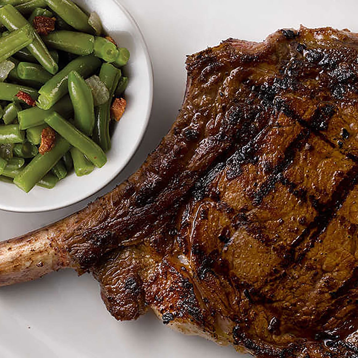 Bone-in steak on an oval white plate next to a small dish of green beans.